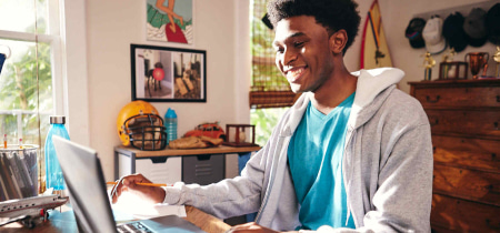 A young man sits at a desk, focused on his laptop, surrounded by a tidy workspace and natural light.