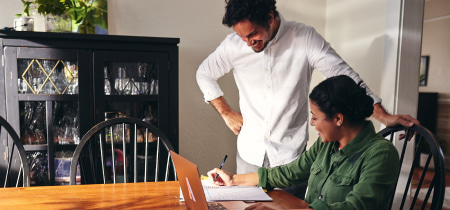 A man and woman collaborate on a laptop at a dining table, focused on their work in a cozy setting.