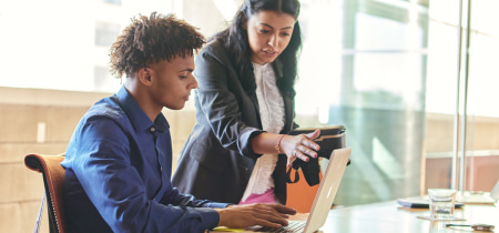 A woman and a man collaborate on a laptop in a modern office setting, focused on their work and sharing ideas.