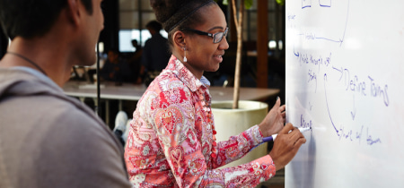 A woman writes on a whiteboard while a man stands attentively behind her, observing the discussion.