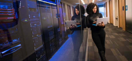 In a hallway, a woman hold a laptop as she engages in conversation and collaborate.