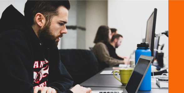 A man in a hoodie is focused on his laptop, engaged in work in a casual setting.