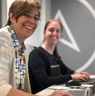 Two women engaged in work, seated at a desk with laptops, collaborating on a project in a professional setting.