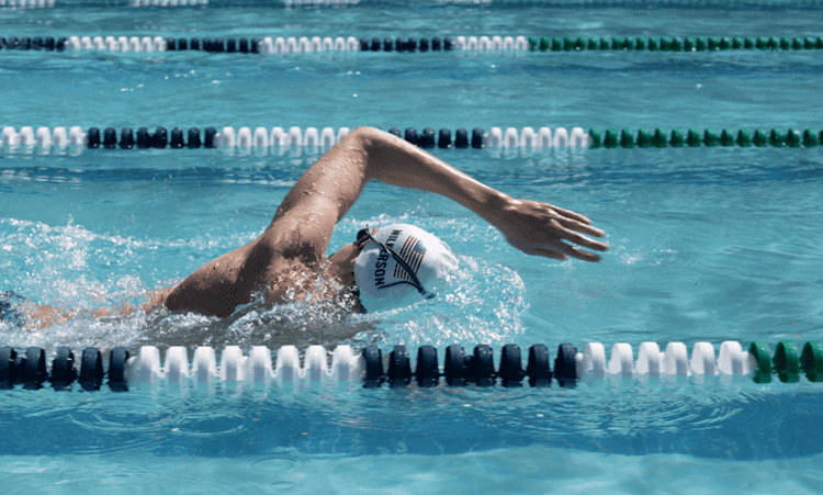 Swimmer in goggles and swim cap in a lane of a clear blue pool, performing freestyle stroke with arm extended forward, conveying focus and effort.